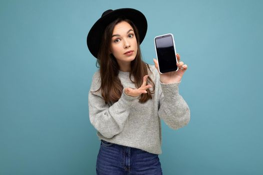 Beautiful young asking dissatisfied brunette woman wearing black hat and grey sweater holding smartphone looking at camera isolated on background. Mock up, cutout