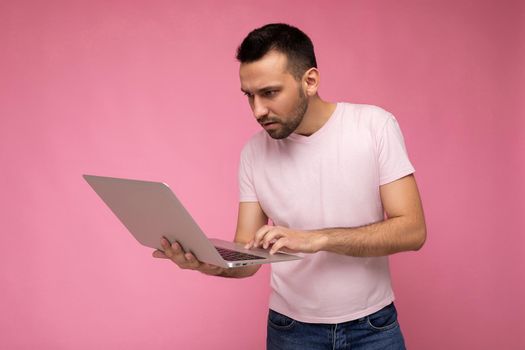 Handsome brunet man holding laptop computer and typing on the keyboard looking at netbook monitor in t-shirt on isolated pink background.