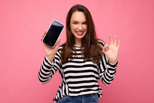 Funny beautiful happy young woman wearing striped sweater isolated over background with copy space showing ok gesture looking at camera showing mobile phone screen display. Mock up, cutout