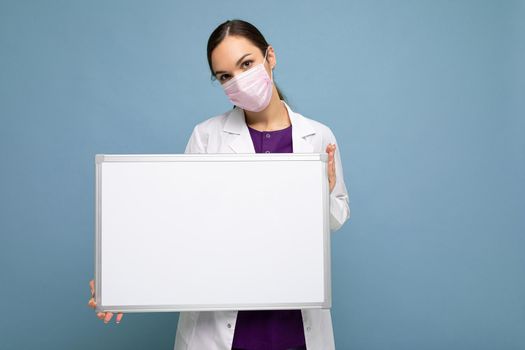 Young attractive nurse in protective face mask and white medical coat holding an empty magnetic board isolated on blue background.