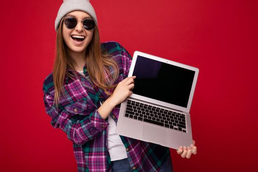 Photo shot of amazing funny happy beautiful smiling dark blond young woman holding computer laptop with empty monitor screen with mock up and copy space wearing sun glasses hat and colourful shirt looking at camera isolated over red wall background.