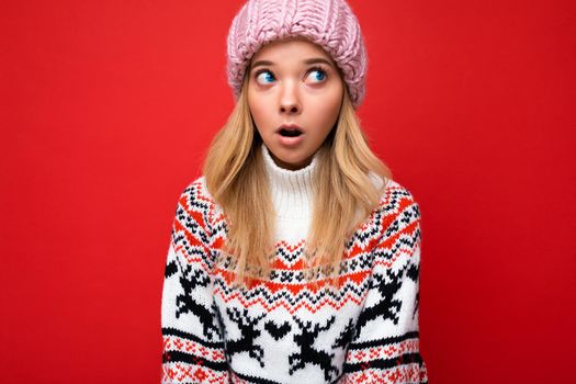 Shot of beautiful astonished young blonde woman isolated over red background wall wearing winter sweater and pink hat looking up to the side.