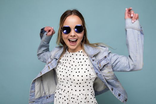 Photo shot of beautiful positive young blonde woman wearing summer casual clothes and stylish sunglasses isolated over colorful background looking at camera and having fun.