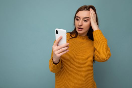 Photo of beautiful shocked young woman wearing orange sweater poising isolated on blue background with empty space holding in hand and using mobile phone communicating with friends looking at gadjet screen.