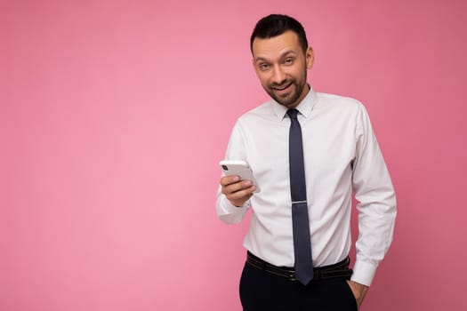 Photo shot of handsome positive good looking adult businessman wearing casual stylish outfit poising isolated on background with empty space holding in hand and using mobile phone messaging sms looking at camera.