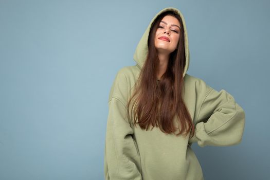Young beautiful woman looking at camera. Trendy girl in casual hipster hoodie clothes. Positive female shows facial emotions. Funny model isolated on blue background with free space for text.