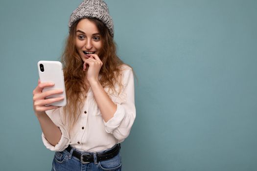 photo of attractive amazed positive young blonde curly woman wearing casual white shirt and grey hat isolated over blue background wall holding phone and using communicating online looking at gadjet display. copy space