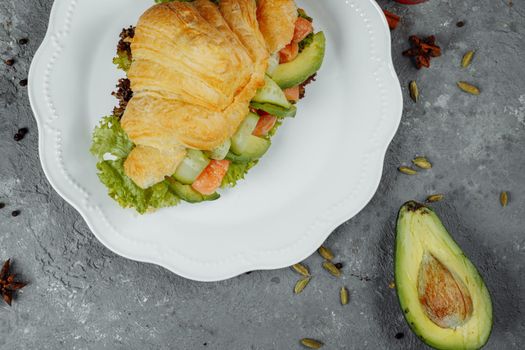 Croissant sandwich with red fish, avocado, fresh vegetables and arugula on black shale board over black stone background. Healthy food concept.