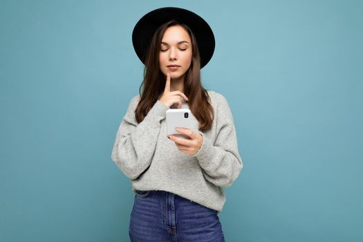 Beautiful young brunette woman wearing black hat and grey sweater holding smartphone looking down at phone texting isolated on background and thinking.