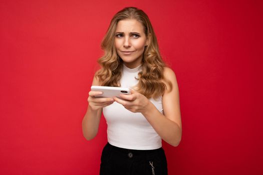 Photo of concentrated beautiful young blonde woman wearing white t-shirt isolated on red background using smartphone and playing phone games looking to the side.