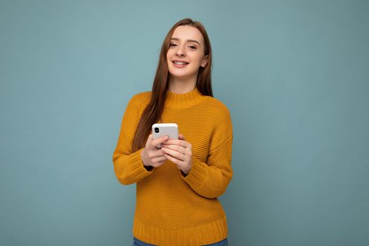 Photo shot of attractive positive good looking young woman wearing casual stylish outfit poising isolated on background with empty space holding in hand and using mobile phone messaging sms looking at camera.