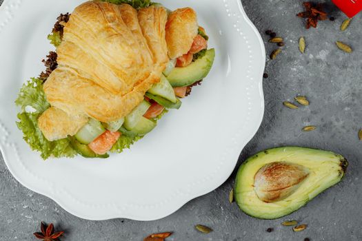 Croissant sandwich with red fish, avocado, fresh vegetables and arugula on black shale board over black stone background. Healthy food concept.