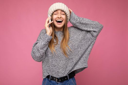 Beautiful positive happy young blonde woman wearing casual grey sweater and beige hat isolated over pink background holding in hand and talking on mobile phone with close eyes.