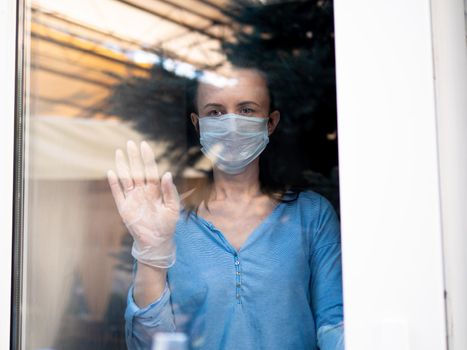 Woman in face virus mask standing behind window glass pane, touching it with hand, looking sad. Woman in a medical mask behind a glass window on self-isolation during an epidemic.