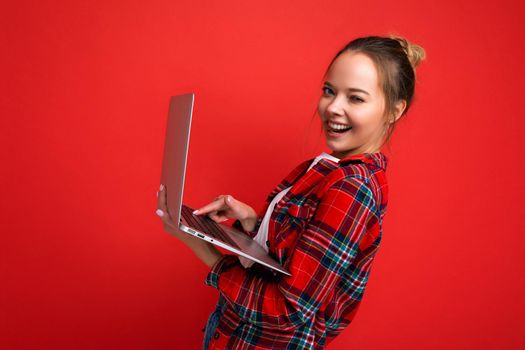 side profile photo of pretty laughing young woman holding netbook looking at camera typing on keyboard wearing red shirt isolated over red wall background.