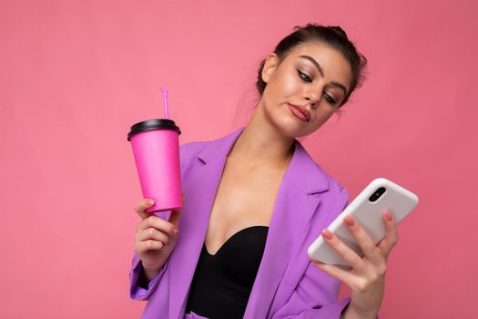 Shot of dissatisfied sad upset caucasian brunette woman holds take away coffee dressed in casual clothes isolated over pink wall uses smartphone. Negative concept.