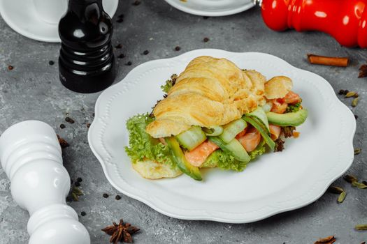 Croissant sandwich with red fish, avocado, fresh vegetables and arugula on black shale board over black stone background. Healthy food concept.