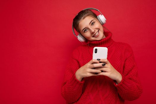 Photo of beautiful joyful smiling young woman wearing stylish casual clothes isolated over background wall holding and using mobile phone wearing white bluetooth headphones listening to music and having fun looking at camera.