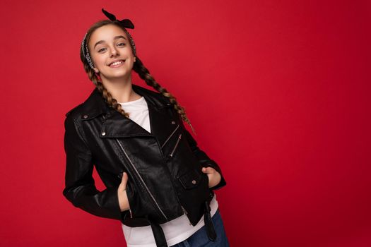 Shot of attractive cool positive smiling brunette little female teenager with pigtails wearing stylish black leather jacket and white t-shirt isolated on red background looking at camera. Empty space, copy space