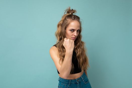 Young dissatisfied attractive winsome blonde curly woman with sincere emotions wearing trendy black top isolated over blue background with free space and showing fist. Negative concept.