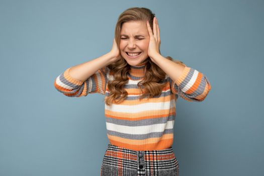 Young emotional beautiful blond curly woman with sincere emotions wearing casual striped sweater isolated on blue background with copy space and covering ears trying not to hear.