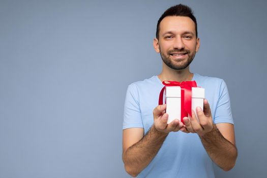 Photo shot of handsome positive smiling brunette unshaven young man isolated over blue background wall wearing blue t-shirt holding white gift box with red ribbon and looking at camera. Empty space