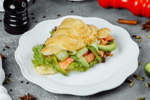 Croissant sandwich with red fish, avocado, fresh vegetables and arugula on black shale board over black stone background. Healthy food concept.