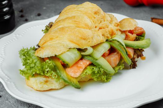 Croissant sandwich with red fish, avocado, fresh vegetables and arugula on black shale board over black stone background. Healthy food concept.