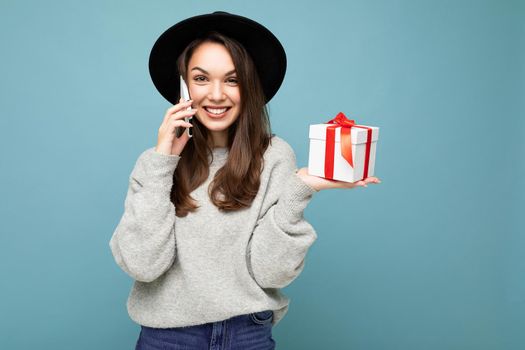 Shot of pretty smiling positive young brunette woman isolated over blue background wall wearing stylish black hat and grey sweater holding gift box talking on smartphone and looking at camera.