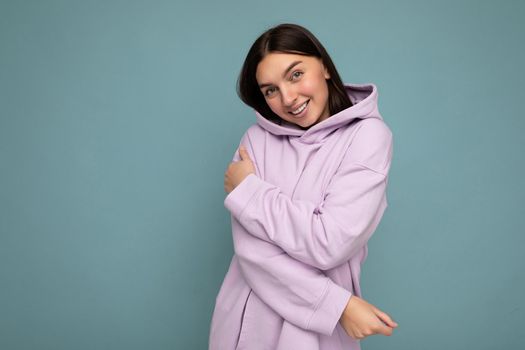 Smiling cute young beautiful brunette woman with sincere emotions isolated on background wall with copy space wearing casual stylish purple hoodie. Positive concept.
