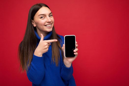 Photo of pretty happy young brunette woman wearing casual blue sweater isolated over red background with empty space holding in hand mobile phone and showing smartphone with empty screen for mockup looking at camera and pointing finger at device.