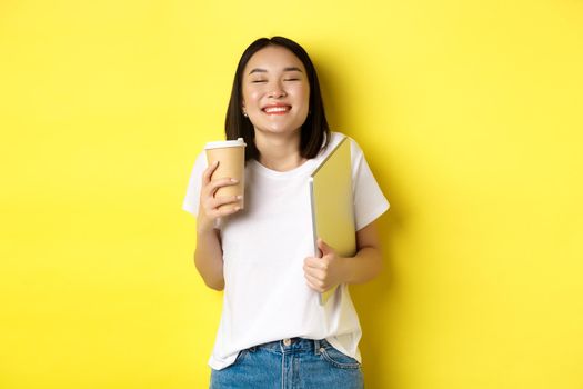 Happy young woman enjoying coffee from cafe and smiling, holding cup and laptop, standing over yellow background.