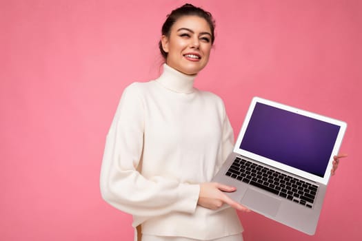 Photo of beautiful brunet young woman with gathered dark hair wearing white sweater holding computer laptop and looking to the side with close eyes and open mouth isolated on pink wall background