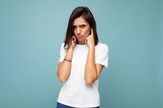 Portrait of touchu sad sorrowful young beautiful brunette woman with sincere emotions wearing casual white t-shirt for mockup isolated on blue background with empty space and covering ears.