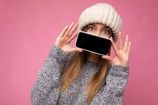 Closeup photo of Beautiful young blonde woman wearing casual grey sweater and beige hat isolated over pink background holding in hand and showing mobile phone with empty display for mockup looking at camera.