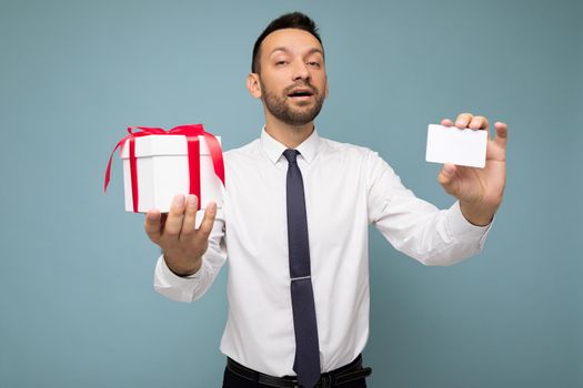 Photo shot of handsome happy brunette young unshaven businessman with beard isolated over blue background wall wearing white shirt and tie holding white gift box with red ribbon and credit card looking at camera.