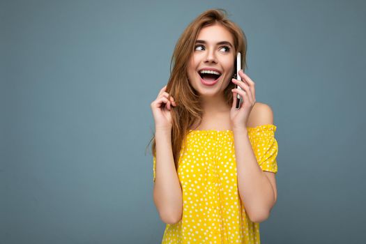 Attractive positive sexy smiling young blonde woman wearing stylish yellow summer dress standing isolated over blue background holding and talking on mobile phone looking to the side. copy space