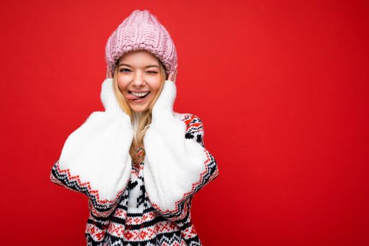 Photo of beautiful happy funny joyful young blonde woman isolated over red background wall wearing winter warm sweater and pink hat looking at camera winking and showing tongue. Free space, copy space