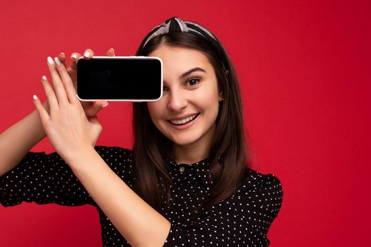 Photo of beautiful smiling girl good looking wearing casual stylish outfit standing isolated on background with copy space holding smartphone showing phone in hand with empty screen display for mockup looking at camera.