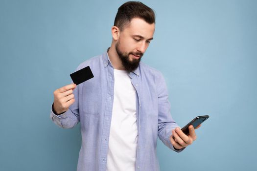 closeup Photo of good looking attractive smiling brunet unshaven young man wearing casual blue shirt and white t-shirt isolated over blue background wall holding credit card and using mobile phone making payment looking at smartphone display.