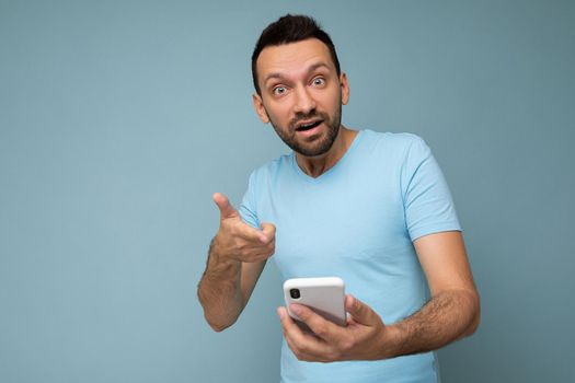 Photo of amazed asking handsome young man with beard wearing everyday blue t-shirt isolated over blue background holding and using mobile phone communication online on the internet looking at camera.