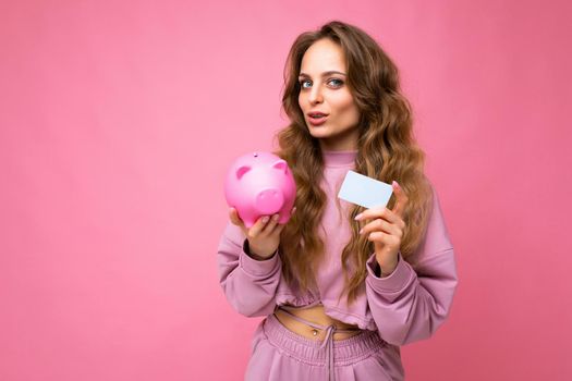 Portrait photo of happy positive young beautiful attractive woman with wavy long blonde hair with sincere emotions wearing stylish pink hoodie isolated over pink background with copy space, holds pink piggy box and credit card. Money concept.