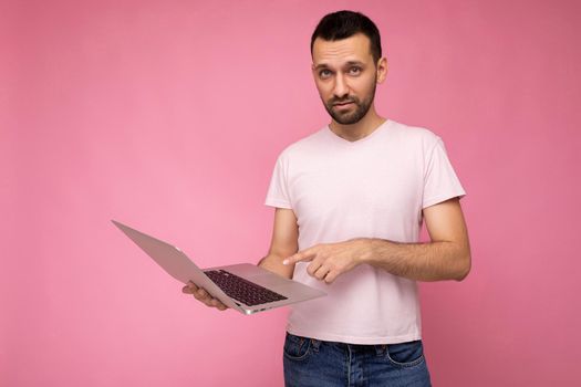 Handsome brunet man holding laptop computer looking at camera and pointing to the screen in t-shirt on isolated pink background.