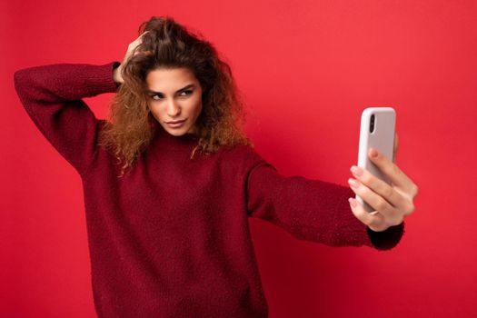Sexy Attractive young blogger woman with curly hair wearing dark red sweater isolated on red background wall holding and using smart phone looking at telephone screen and taking selfie.