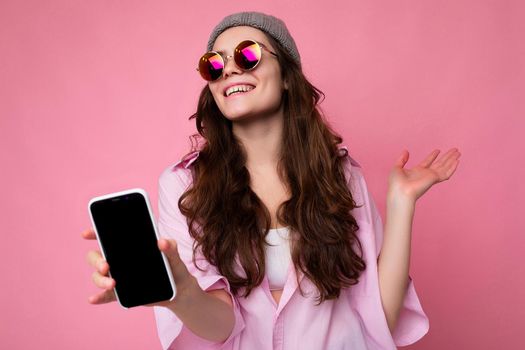 Positive smiling attractive young brunette woman wearing stylish pink shirt grey hat and colourful sunglasses isolated over pink background holding in hand and showing mobile phone with empty display for mockup looking at camera
