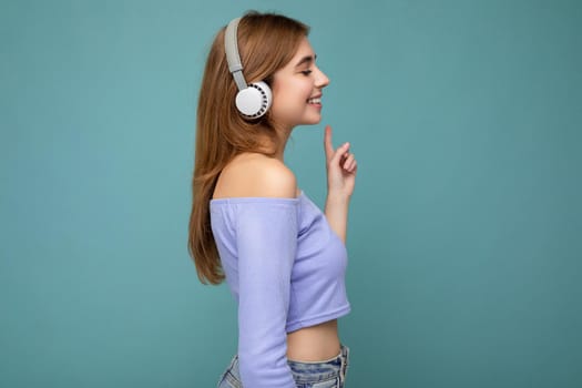 Side-profile photo of beautiful positive smiling young blonde woman wearing blue crop top isolated over blue background wall wearing white wireless bluetooth earphones listening to cool music and looking to the side.