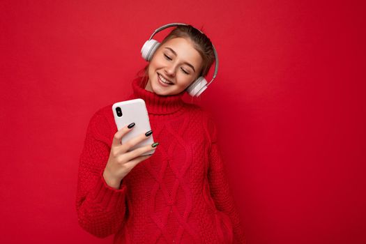 Photo shot of beautiful joyful smiling young female person wearing stylish casual outfit isolated over colorful background wall wearing white bluetooth wireless earphones and listening to music and using mobile phone looking at gadjet display.