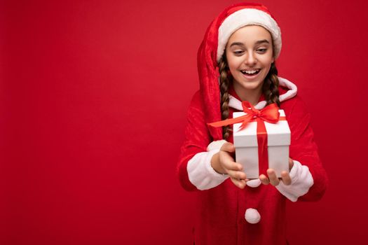 Shot of beautiful happy smiling brunette girl isolated over red background wall wearing new year santa claus outfit holding white gift box with red ribbon. Empty space