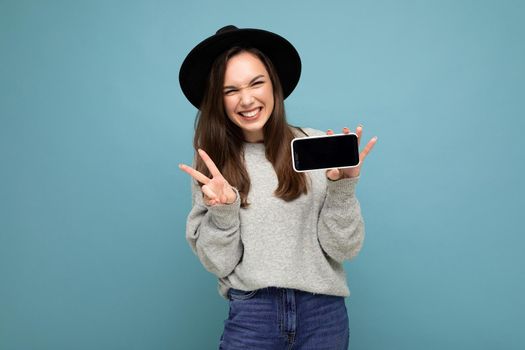 Pretty positive young female person wearing black hat and grey sweater holding phone showing smartphone isolated on background showing peace gesture.