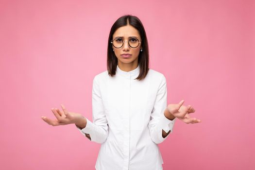 Attractive asking dissatisfied young brunette woman wearing everyday stylish clothes and modern optical glasses isolated on colorful background wall looking at camera and having doubts.